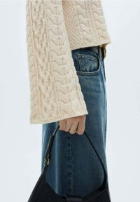 Pull en tricot beige à mailles larges, associé à un jean en denim bleu taille haute, tenant un sac à main noir avec une poignée courbée.