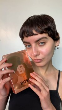 Scatola rettangolare color oro rosa con un design circolare, con la parola "BOSS" in testo nero e grassetto. Mani che tengono la scatola. Superficie dalla texture liscia.