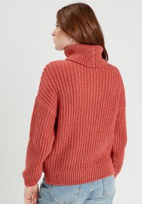 Pull en tricot couleur rouille avec un col montant, motif en relief texturé et épaules tombantes. Coupe légèrement oversized et poignets côtelés.