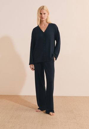 Donna bionda in piedi a piedi nudi che indossa una maglia a coste blu scuro a maniche lunghe con bottoni e pantaloni larghi abbinati su pavimento e muro beige.