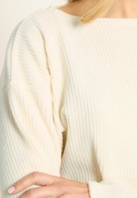 Cremfarbener Rippstrickpullover aus weichem Stoff, mit weitem Ausschnitt und fallenden Schulternähten. Sichtbare Struktur mit vertikalen Streifen.