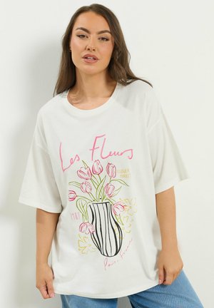 Kobieta w białej, oversize'owej koszulce z różowymi tulipanami w paskowanym wazonie oraz napisem „Les Fleurs”, „Flower Market”, „Paris, France”.