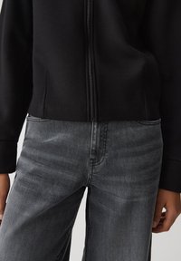 Giacca nera con zip e finitura testurizzata, abbinata a jeans grigi scuri a gamba larga con una leggera sfumatura e cuciture visibili.