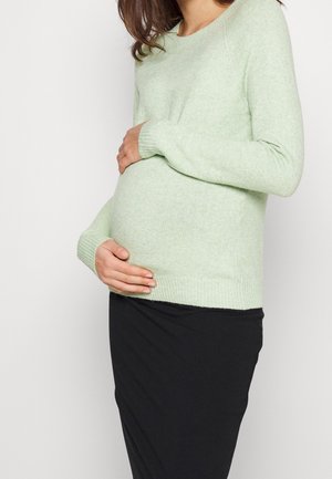 Těhotná osoba v světle zeleném svetru si oběma rukama jemně objímá břicho na jednoduchém světlém pozadí.
