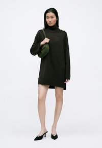 Καφέ πλεκτό φόρεμα με ψηλό λαιμό και ριμπέ σχέδιο στο τελείωμα και κουμπιά στους ώμους. Συνδυασμένο με πράσινη τσάντα χιαστί και μαύρες μυτερές γόβες.