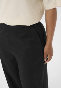 Persoon in zwarte op maat gemaakte broek en een crèmekleurige top met korte mouwen, voorzien van kleine decoratieve studs, staand tegen een effen achtergrond.