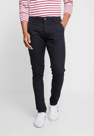 Persoon die zwarte slim-fit broek draagt, witte sneakers en een wit shirt met lange mouwen met rode horizontale strepen, staand tegen een effen achtergrond.