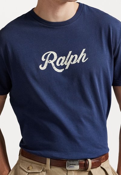 Tengerészkék pamut póló, amelyen egy megkopott fehér "Ralph" grafika található a mellkasán, khaki nadrággal és barna övvel párosítva.