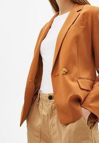 Blazer orange avec une fermeture à un seul bouton, des revers crantés et deux poches avant. Associé à un haut blanc et un pantalon beige.