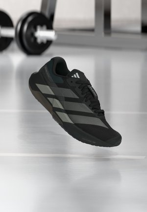 Zwart-grijze sportschoen met Adidas-strepen die zweeft op een sportschoolvloer, met gewichthefapparatuur wazig op de achtergrond.