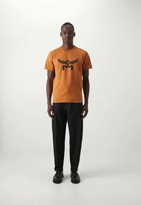 Mann som står iført en oransje T-skjorte med et svart grafisk motiv, svarte bukser og svarte sko mot en enkel lys bakgrunn.