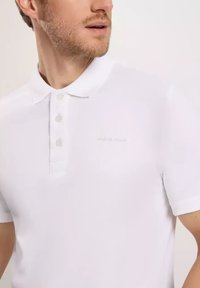 Biała koszulka polo z teksturowanej tkaniny, z klasycznym kołnierzykiem, trzyguzkowym zapięciem oraz subtelnym logiem marki na piersi.