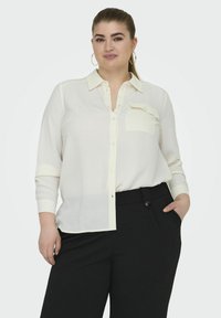 Cămașă albă cu nasturi, din material texturat, cu mânecă lungă, un buzunar pe piept și croială lejeră. Asortată cu pantaloni negri, ajustați.