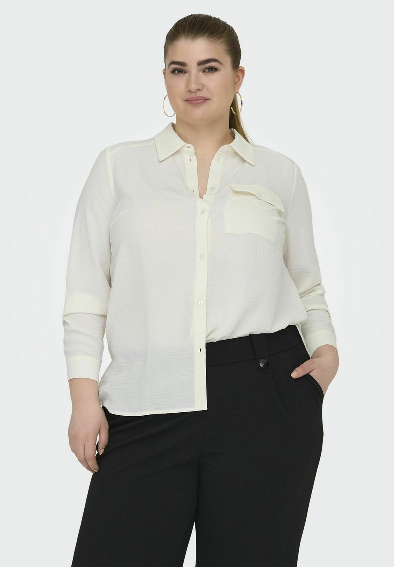 Cămașă albă cu nasturi, din material texturat, cu mânecă lungă, un buzunar pe piept și croială lejeră. Asortată cu pantaloni negri, ajustați.