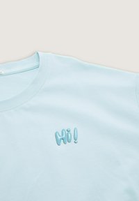 Camiseta de manga corta color azul claro con un escote redondeado y un detalle bordado texturizado de "¡Hola!" en el frente. Tejido suave, corte relajado.