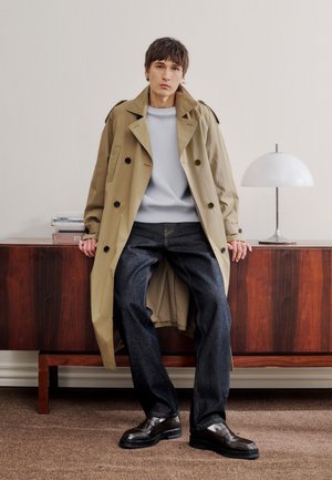 Joven con abrigo beige, suéter gris claro, vaqueros oscuros y zapatos negros, sentado en un aparador de madera junto a una lámpara de mesa blanca.