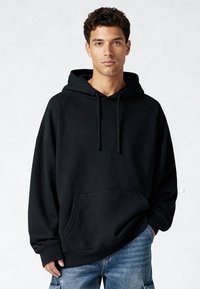 Zwarte oversized hoodie met een koord, een kangaroozak aan de voorkant en geribbelde manchetten, gedragen met lichtblauwe denim shorts. Gladde textuur.