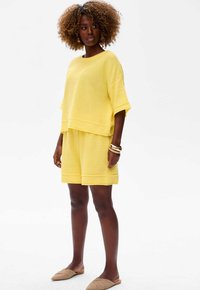 Żółty zestaw odzieży domowej z dzianiny, składający się z koszulki z krótkim rękawem i szortów, o luźnym kroju i prążkowanych akcentach; uzupełniony beżowymi butami wsuwanymi.