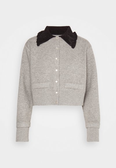 Pilkas trumpas kardiganas, pagamintas iš minkštos medžiagos, su juodu raukšlėtu apykakle. Jame yra mygtukų užsegimai ir dvi priekinės kišenės.