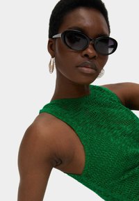 Grünes gestricktes Tanktop mit strukturierter Oberfläche, breitem Ausschnitt und offenem Schulterdesign. Schwarze ovale Sonnenbrille und goldene Creolen.