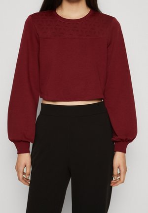 Vrouw draagt een cropped, bordeauxrode top met lange mouwen en een getextureerde inzet met ballonmouwen, gecombineerd met een hooggetailleerde zwarte broek.