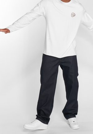 Homme portant une chemise blanche à manches longues avec un petit motif graphique et un jean large foncé, debout, les bras étendus devant un fond blanc.