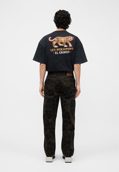 Άνδρας με πλάτη, φοράει μαύρο υπερμεγέθη T-shirt με ροζ και κίτρινο μοτίβο λεοπάρδαλης και κείμενο «Les Benjamins El Gringo», μαύρο παντελόνι με σχέδια και πλατφόρμες.