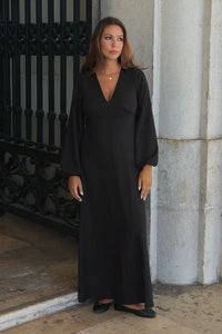 Abito lungo nero a maniche lunghe con scollatura a V profonda, corpo arricciato e silhouette fluida. Indossato con scarpe nere basse, davanti a un muro di pietra.
