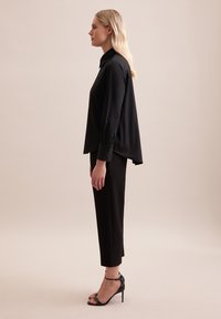 Schwarze Bluse mit langen Ärmeln und Kragen, lockere Passform und Falten im Rücken. Kombiniert mit schmalen schwarzen Hosen und schwarzen High-Heel-Sandalen.