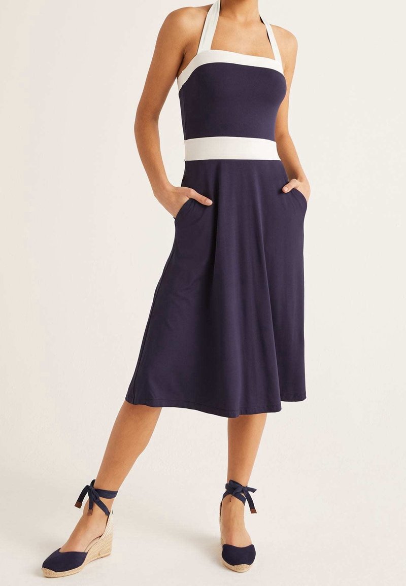 Femme portant une robe midi bleu marine sans manches avec des bretelles halter blanches et une ceinture à la taille, associée à des espadrilles compensées bleu marine avec des liens à la cheville.