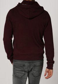 Burgunderfärgad huvtröja med långa ärmar, ribbade muddarna och midjan, visad bakifrån. Bärs tillsammans med grå jeans med bakfickor.