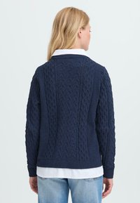 Marineblauer Strickpullover mit strukturierten Zopfmuster, geripptem Kragen und Bündchen sowie einer lässigen Passform, über einem weißen Hemd getragen.