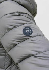 Siva jaja jakna s rukavima koja ima dijagonalno prešane dijelove i okrugli logotip u tamnoplavoj boji s bijelim tekstom.