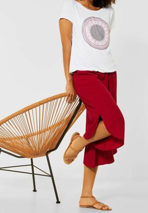 Witte t-shirt met een ronde roze grafische design, gecombineerd met rode wijde broek en bruine sandalen met bandjes, tegen een minimalistische achtergrond.