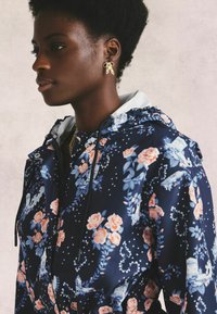 Floral-patterned navy jacket features a hood, a zipper, and drawstrings. Gold earrings add contrast. Smooth fabric with a glossy finish.