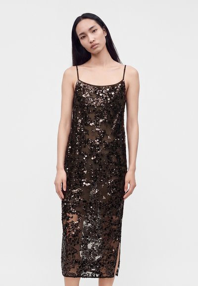 Femme aux longs cheveux noirs portant une robe midi noire sans manches à sequins avec fines bretelles, debout devant un fond clair uni.
