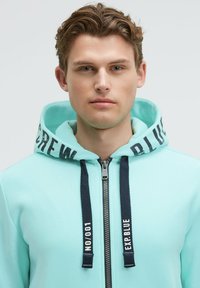Felpa con zip verde menta, dotata di lacci blu navy, con la scritta "EXP. BLUE" sulla cappuccio. Tessuto morbido e vestibilità standard.