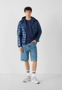 Marineblauwe pufferjack met een glanzende afwerking, ritssluiting aan de voorkant en capuchon; gecombineerd met een marineblauw shirt, denim shorts en grijze sneakers met witte sokken.
