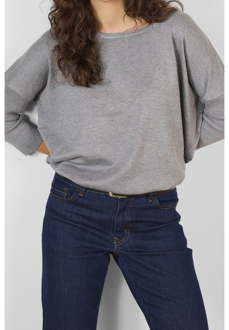 Femme portant un pull en maille gris ample et un jean bleu foncé, les mains sur les hanches, sur un fond clair uni.