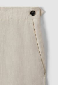 Pantalons en lin beiges avec un unique bouton noir, des poches latérales et une surface en tissu texturé. Détail de taille repliée visible.