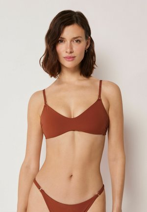 Frau mit schulterlangem dunklem Haar, die ein rostfarbenes Bikinioberteil und passende Unterteile vor einem schlichten Hintergrund trägt.