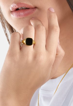 Hand with gold ring featuring a black square stone near lips, wearing white top and gold chain necklace.