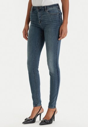 Persona che indossa jeans blu aderenti e scarpe slingback nere con tacchi alti in piedi davanti a uno sfondo bianco.