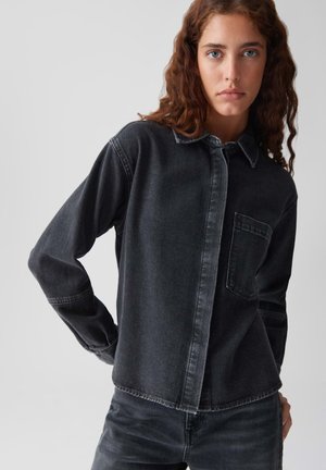Jonge vrouw met krullend bruin haar die een zwarte denim blouse en bijpassende zwarte denim broek draagt voor een effen achtergrond.