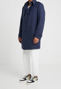 Cappotto di lana blu navy con design doppiopetto, caratterizzato da grandi bottoni, tasche laterali e polsini arrotolati, indossato sopra pantaloni bianchi e sneakers.