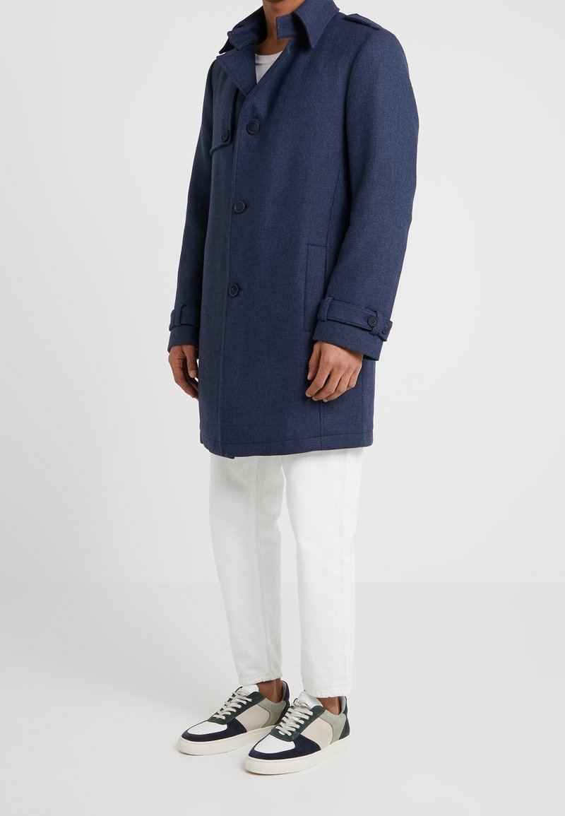 Cappotto di lana blu navy con design doppiopetto, caratterizzato da grandi bottoni, tasche laterali e polsini arrotolati, indossato sopra pantaloni bianchi e sneakers.