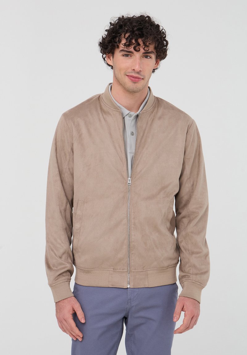 Giacca bomber color noce realizzata in morbido tessuto scamosciato, con zip frontale, orlo e polsini a costine, e tasche laterali. Indossata sopra un polo grigio chiaro.