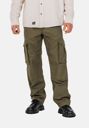 Hombre con pantalones cargo verde oliva con bolsillos laterales, zapatos negros gruesos y una camisa beige abotonada, de pie contra un fondo liso.
