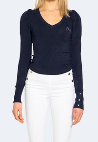 Femme portant un pull bleu marine ajusté à col en V avec des épaules bouffantes et des boutons argentés sur les poignets, associé à un pantalon blanc taille haute.