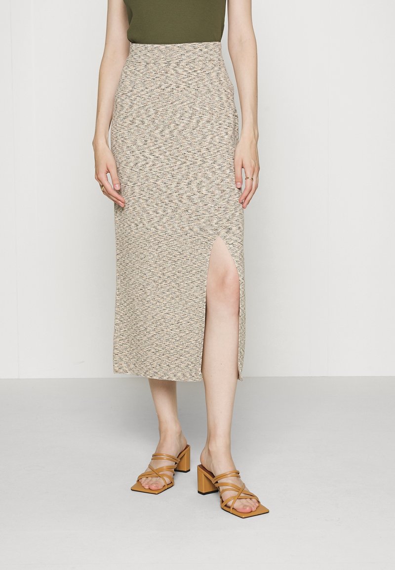 Kvinde iført beige tekstureret midinederdel med frontslids og tanfarvede rem-sandaler med blokhæl, stående på lyst gulv mod hvid væg.